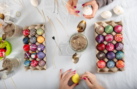 human hands of two sorbian women are painting with melting wax with a trimmed goose quill and a needle head on the surface of the egg at a Table with colorful painted easter eggs, copy space