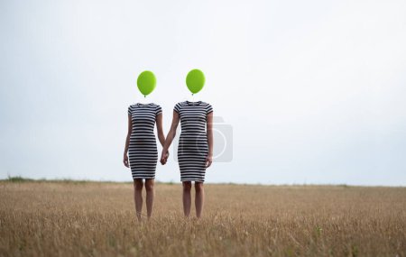 Photo for Two happy sexy young women, in black and white striped dress, friends, holding hands, heads replaced by green balloons stand hand in hand on harvested corn field, copy space - Royalty Free Image