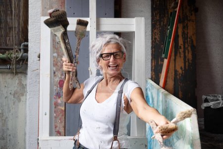 smiling laughing happy older mature woman portrait, proud artist, in her fifties with grey hair and black glasses, with a positive gesture persuasiveness and charisma and many paintbrushes, copy space