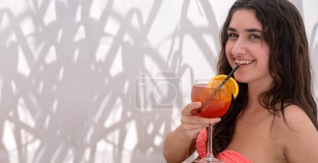 Joyeux sourire jeune femme sexy en bikini avec un cocktail de vodka orange sang dans le salon du bar de la plage, fond de tissu illuminé avec nuance de roseaux, motifs abstraits d'ombre de silhouette, espace de copie