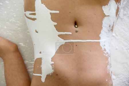 Bauchbild einer nackten Frau mit glänzendem Nabelpiercing, übergossen mit weißer Farbe, bespritzt mit weißer Farbe, Kopierraum