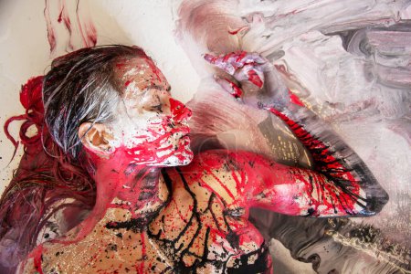 Portrait of sexy nude sensual seductive female woman with long brunette hair, doused with white, red and black paint, lying elegant on the Studio floor, Color reminds of bloody splatter, Copy space