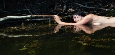 beautiful young sexy nude brunette wet woman mermaid nymph sensual sensitive seductive in green natural water, lake, pond, clung to a branch lying in the water, copy space