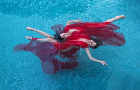 Top view to two beautiful young sexy woman in red dress, towel floats weightlessly elegant water ballet swimming in the water of the turquoise pool, copy space