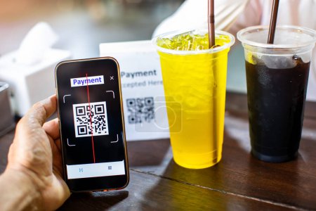 Selektiver Fokus auf die Hand mit dem Smartphone, um QR-Code auf Tag mit verschwommenem Chrysanthemensaft und Eiskaffee im Café zu scannen, um digitales Bezahlen ohne Geld zu generieren. Qr-Code-Bezahlkonzept.