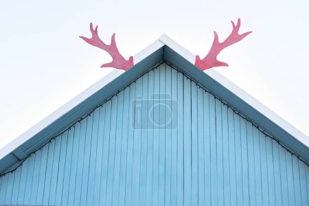 Foto de Decoración de Navidad en el techo. Cuernos rojos festivos decorativos de un reno en una casa de madera contra el cielo. Techo Sod cabaña de caza tradicional. cuernos de ciervo en adjunto en la parte superior del techo de la casa. - Imagen libre de derechos