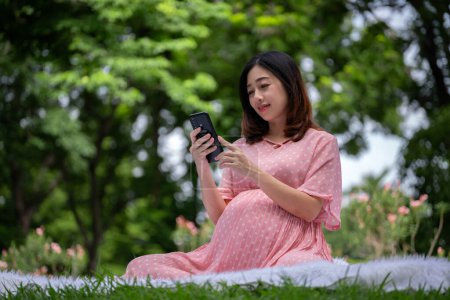 Foto de Retrato de mujer embarazada asiática relajándose y usando smartphone en el parque - Imagen libre de derechos