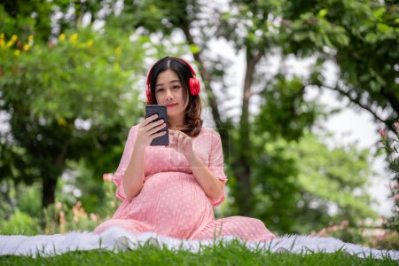 Foto de Retrato de mujer embarazada asiática relajándose con el uso de teléfonos inteligentes y auriculares para escuchar la canción en el parque - Imagen libre de derechos