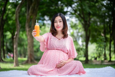 Foto de Retrato de mujer embarazada asiática relajándose y bebiendo jugo de naranja en el parque - Imagen libre de derechos