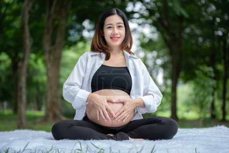 Foto de Retrato de mujer embarazada asiática relajándose y posando con las manos en forma de corazón en el vientre en el parque - Imagen libre de derechos