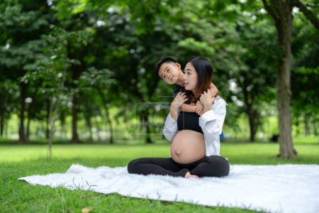 Foto de Retrato de una mujer embarazada relajándose en el parque con su hijo. Pequeño hijo abrazando madre desde detrás embarazada - Imagen libre de derechos