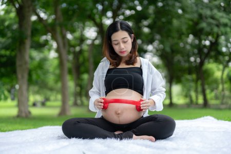 Foto de Retrato de mujer embarazada asiática relajándose y en el parque. Mujer joven usa auriculares en su estómago para dejar que su feto escuche música. - Imagen libre de derechos