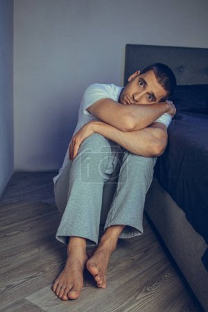 Photo for Dramatic lifestyle portrait of a handsome guy in his 30s and 40s, sitting sadly on the bed, feeling anxious and suffering from depression. Attractive depressed and upset man in home bedroom - Royalty Free Image