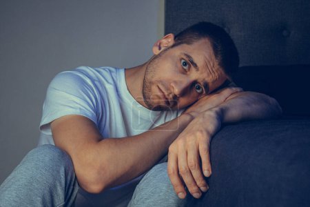 Photo for Dramatic lifestyle portrait of a handsome guy in his 30s and 40s, sitting sadly on the bed, feeling anxious and suffering from depression. Attractive depressed and upset man in home bedroom - Royalty Free Image