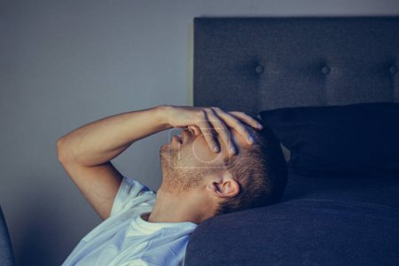 Dramatic lifestyle portrait of a handsome guy in his 30s and 40s, sitting sadly on the bed, feeling anxious and suffering from depression. Attractive depressed and upset man in home bedroom