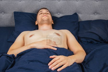 Foto de Handsome young man sleeping comfortably on the bed at night in his bedroom without clothes. Bachelor bedroom. Deep sleep - Imagen libre de derechos