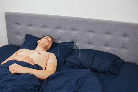 Téléchargez les photos : Handsome young man sleeping comfortably on the bed at night in his bedroom without clothes. Bachelor bedroom. Deep sleep - en image libre de droit