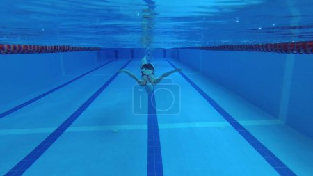 Téléchargez les photos : Nageuse sportive en survêtement, en masque de plongée, en bonnet de bain s'entraîne dans la piscine. L'athlète nage sous l'eau. Prise de vue sous-marine, grand angle, 4k. - en image libre de droit