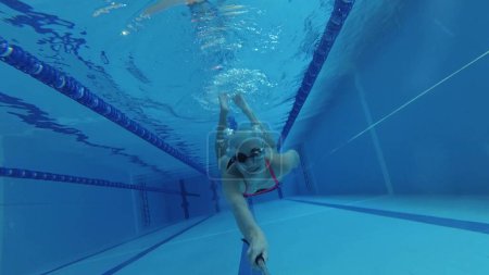 Téléchargez les photos : Nageuse sportive en survêtement, en masque de plongée, en bonnet de bain s'entraîne dans la piscine. Une athlète nage sous l'eau avec une caméra d'action à la main. Prise de vue sous-marine, grand angle, 4k. - en image libre de droit