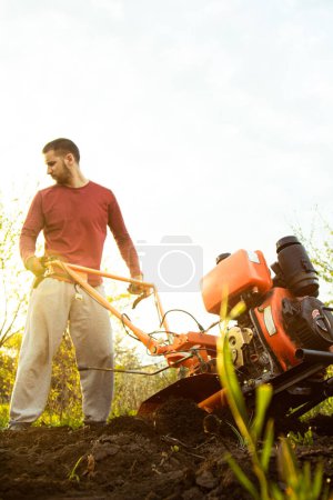 Téléchargez les photos : A farmer man works in the field, plowing the land with a plow on the farm. A plowman on a walk behind a motor cultivator. Tillage season. Organic cultivation of natural products. Agriculture - en image libre de droit