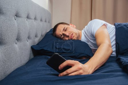 Photo for Happy young cheerful man in pajamas wearing a sleep mask. Rest, rest at home, sit, turn around under a blanket, blanket, point a finger at a mobile phone, isolated on a gray background - Royalty Free Image