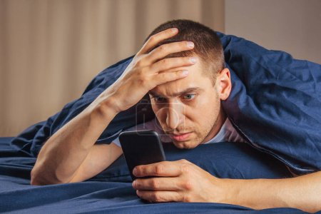 Photo for Bad news, problems, offers and advertising. Shocked upset attractive guy reads a message on the phone and presses his hand to his face on a white bed in the bedroom, free space - Royalty Free Image