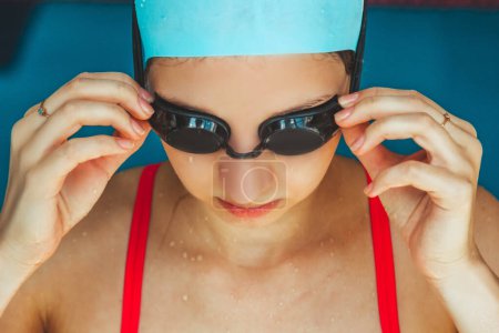 Foto de Extremo primer plano de una cabeza de niña caucásica con una gorra de natación y las manos poniendo gafas de natación, preparándose para el entrenamiento en la piscina - Imagen libre de derechos