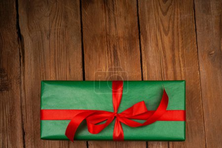 Foto de Caja de regalo verde festivo con lazo de satén rojo, cinta. Lugar para texto, publicidad. Concepto de vacaciones. Un regalo sobre un fondo de madera artesanal. Día de San Valentín, Día de la Mujer, Día de la Madre, Cumpleaños, Boda, Navidad - Imagen libre de derechos