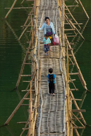 Téléchargez les photos : Luang Prabang, Laos - 15 déc. 2019 : Une femme traverse un pont en bois et en bambou avec ses deux jeunes enfants sur la rivière Nam Khan alors qu'elle traverse Luang Prabang, Laos - en image libre de droit