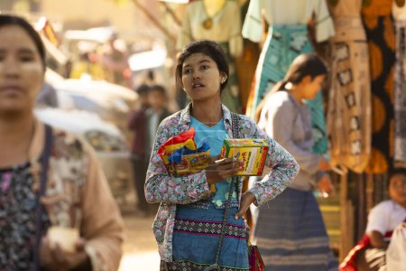 Téléchargez les photos : Bagan, Myanmar - 25 Dec, 2019 : Une jeune fille, avec thanakha sur le visage, marche à travers l'un des marchés de Bagan, avec des sacs et une boîte de riz dans ses mains. - en image libre de droit