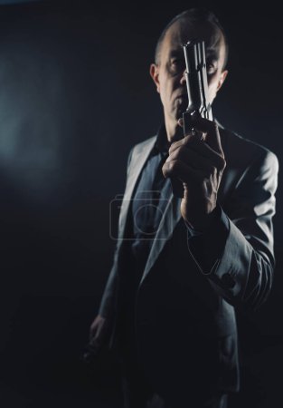 Téléchargez les photos : Espion en costume élégant avec pistolet à pistolet dans le studio sombre photo prise avec fond uni et espace de copie. - en image libre de droit