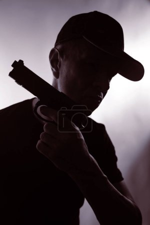 Téléchargez les photos : Jeune tueur criminel en casquette avec pistolet dans la silhouette sombre couverture de livre photo thriller design. - en image libre de droit