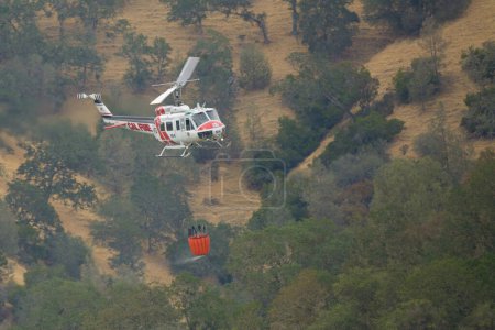 Photo for A Cal Fire Helicopter flying through the hills with a water bucket. Lake Berryessa, California, USA - Royalty Free Image