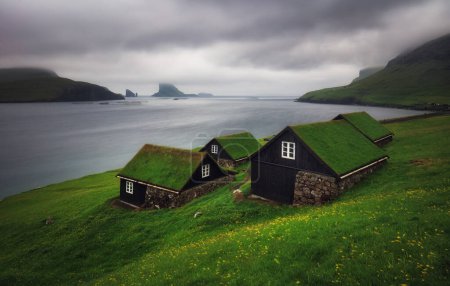 Césped extra faroese - casas cubiertas en el pueblo Bour. Drangarnir y Tindholmur pilas de mar en el fondo. Isla Vagar, Islas Feroe, Dinamarca. Panorama del paisaje