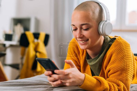 Photo for Close up of teenager bald female with headphones using mobile phone in dorm room - Royalty Free Image