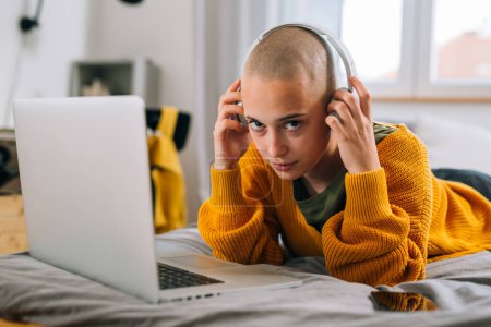 Téléchargez les photos : Jeune femme adulte met des écouteurs tout en utilisant un ordinateur portable dans sa chambre - en image libre de droit