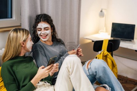 Foto de Amigas con máscara de crema cosmética en la cara disfrutando del tiempo juntos en la habitación - Imagen libre de derechos