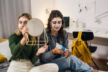 Téléchargez les photos : Adolescent amis appliquer la crème cosmétique sur le visage dans la chambre de dortoir - en image libre de droit