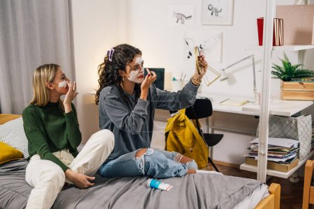 Téléchargez les photos : Adolescent amis appliquer la crème cosmétique sur le visage dans la chambre de dortoir - en image libre de droit