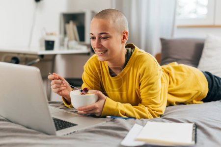 Téléchargez les photos : Adolescente étudiante se détendre dans sa chambre en utilisant un ordinateur portable et manger de la salade de fruits - en image libre de droit