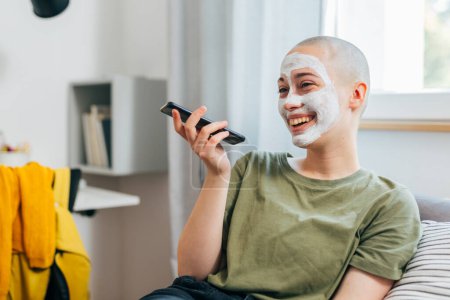 Photo for Teenager female shaved head with cosmetic face cream talking on mobile phone in her room - Royalty Free Image