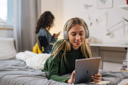 Foto de Adolescente caucásico hembra con auriculares usando tableta digital mientras está acostado en la cama en el dormitorio - Imagen libre de derechos