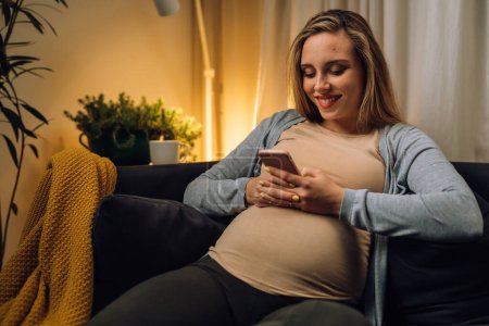 Téléchargez les photos : Femme enceinte s'assoit sur le canapé et regarde le téléphone - en image libre de droit