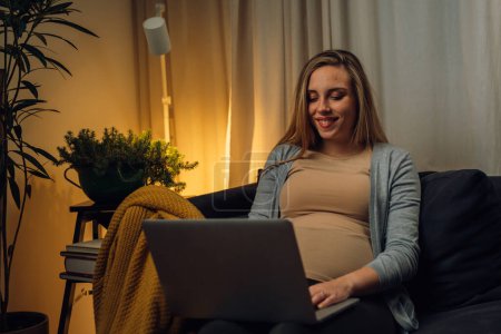 Téléchargez les photos : La femme enceinte regarde son ordinateur portable dans un salon confortable - en image libre de droit