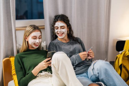 Téléchargez les photos : Deux jeunes femmes portant des masques s'assoient sur le lit et regardent le téléphone - en image libre de droit