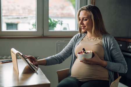 Foto de Hermosa mujer embarazada tiene frutas para un aperitivo mientras mira la tableta - Imagen libre de derechos
