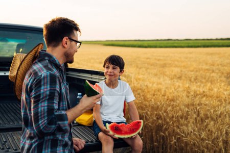 Téléchargez les photos : Père et fils s'amusent dans les champs tout en mangeant des tranches de pastèque - en image libre de droit
