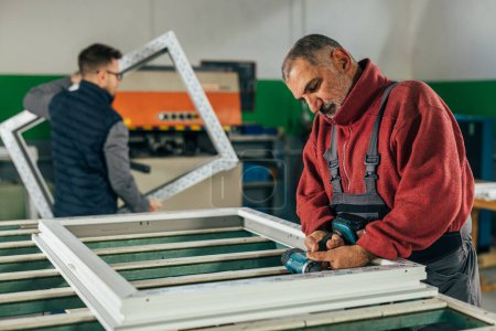 Téléchargez les photos : Travailleur mature visse le cadre de fenêtre en PVC dans l'atelier - en image libre de droit