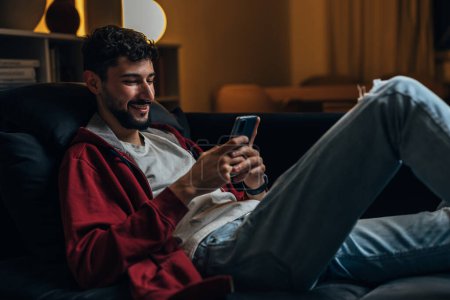 Photo for Man sits on the sofa in the living room and uses his smart phone - Royalty Free Image