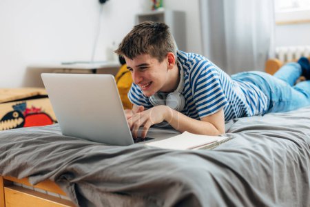 Téléchargez les photos : Teenage boy is laying on his bed and using his laptop - en image libre de droit